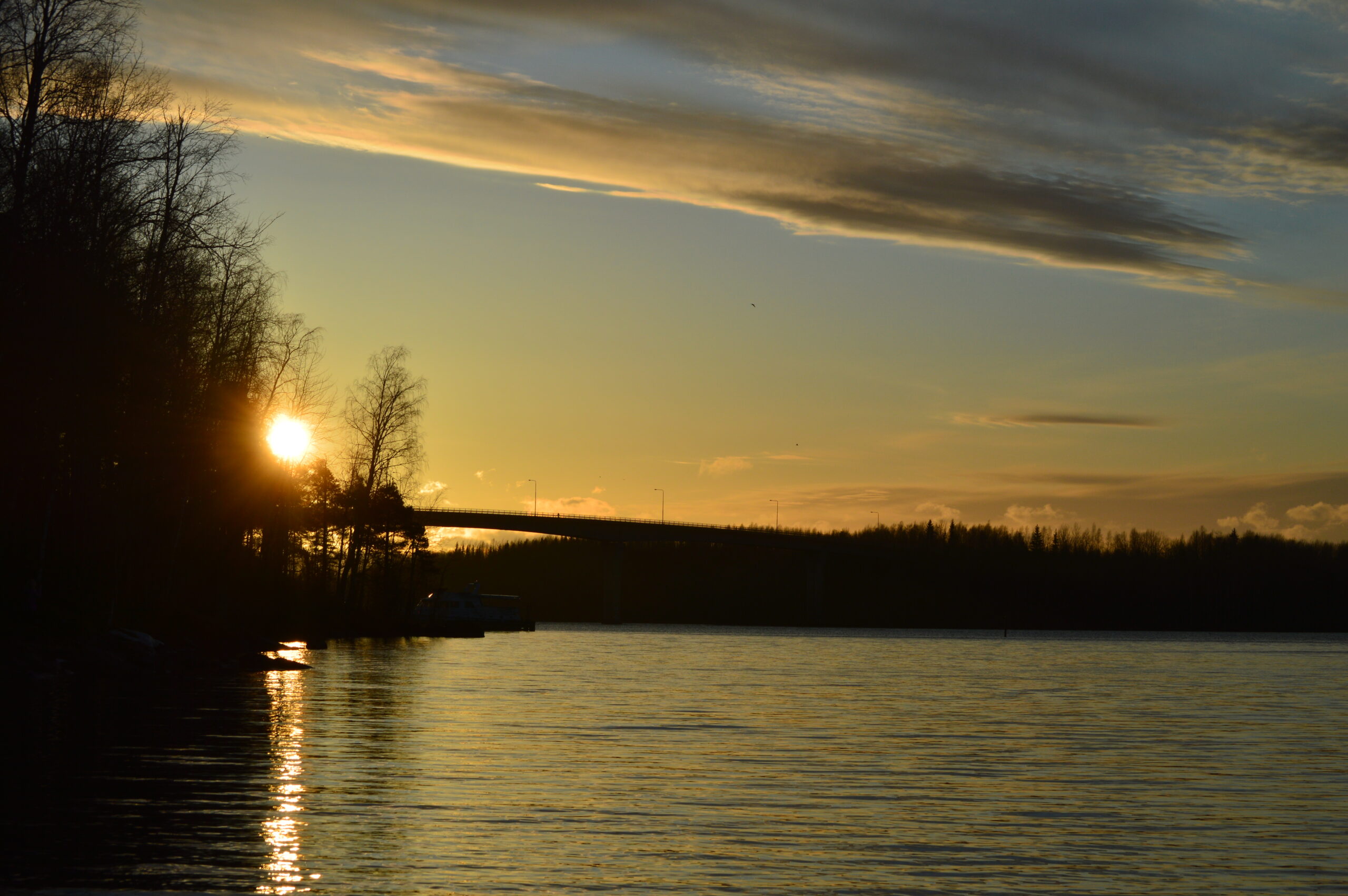 Aurinkolasku kuva Saimaalta. Kuvasssa järven selkää, taivaalla punertavia pilviä. Taustalla sillan musta silhuetti.