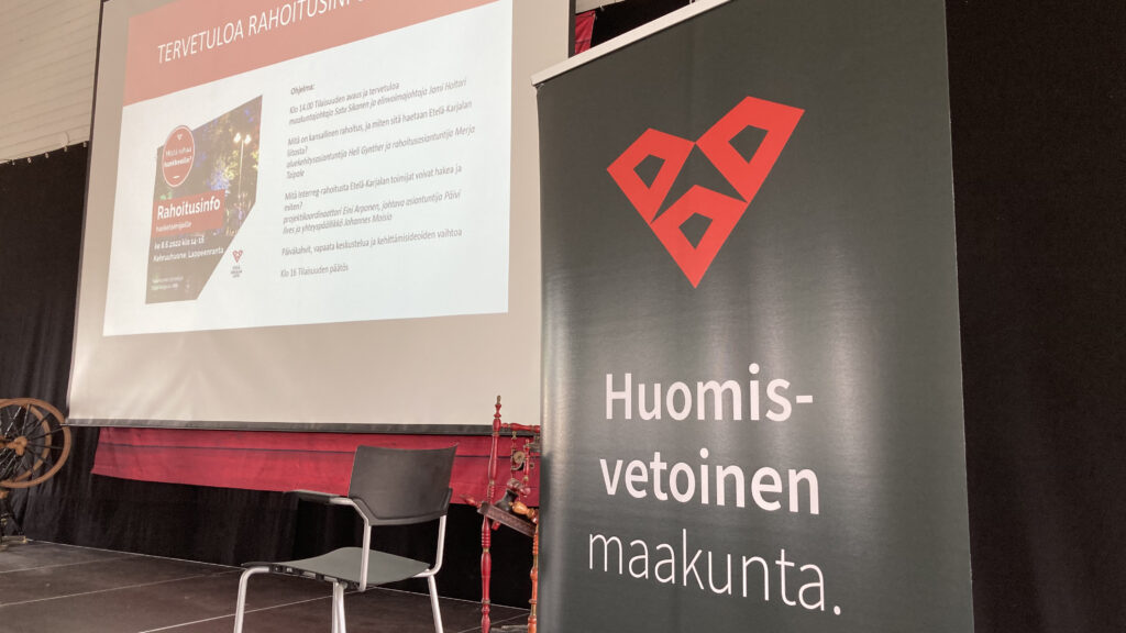 Etualalla musta roll up, jossa on Etelä-Karjalan punainen sydäntunnus ja valkoinen teksti "Huomisvetoinen maakunta". Takana näkyy valkokankaalla rahoitusinfotilaisuuden ohjelma.