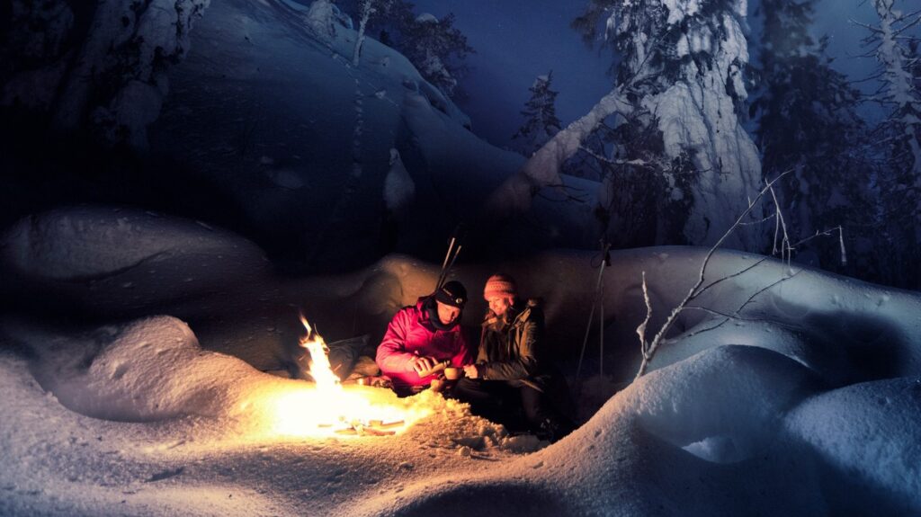 Kuvassa talvisessa metsässä nuotionäärellä kaksi retkeilijää nauttimassa kuumaa juotavaa.