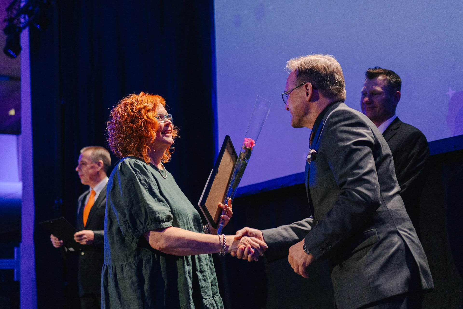 Punahiuksinen nainen vastaanottaa ruusun ja kunniakirjan mieheltä.